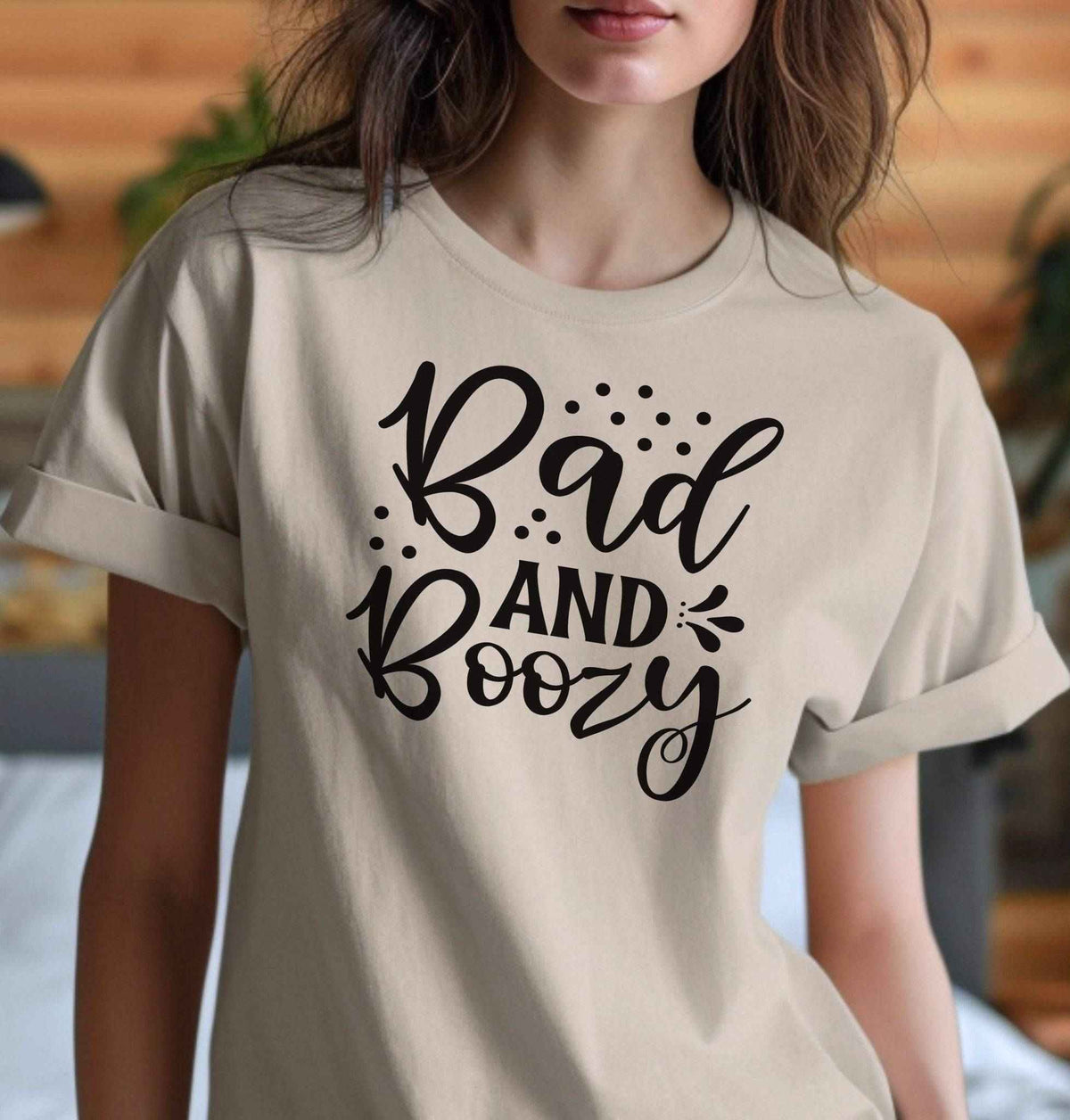 Woman wearing a sand colored Bad and Boozy graphic T Shirt featuring playful black script lettering and decorative accents, perfect for girls' nights and parties.