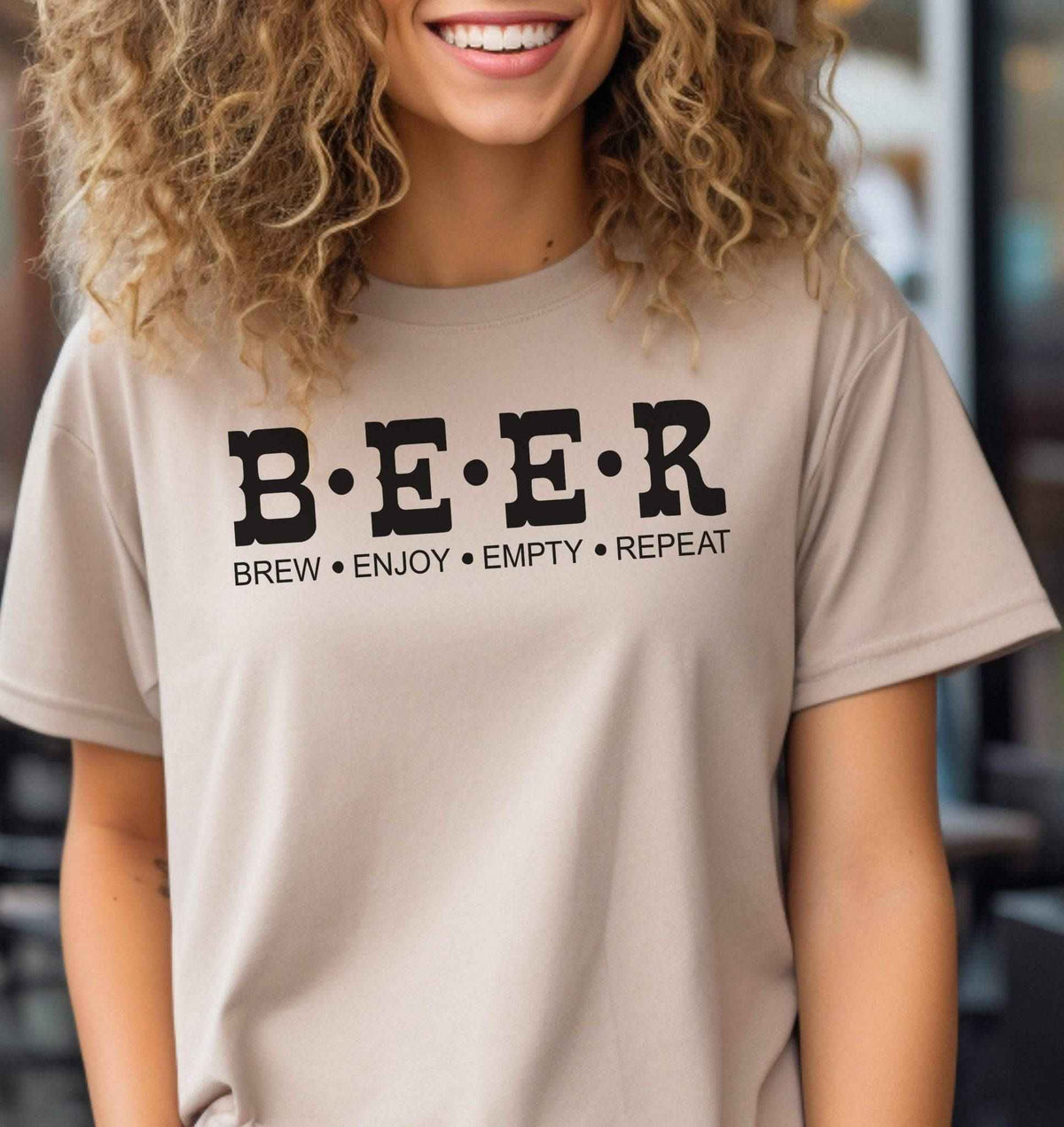 Woman wearing a sand colored B.E.E.R. graphic T Shirt featuring bold black lettering and the phrase Brew Enjoy Empty Repeat underneath.