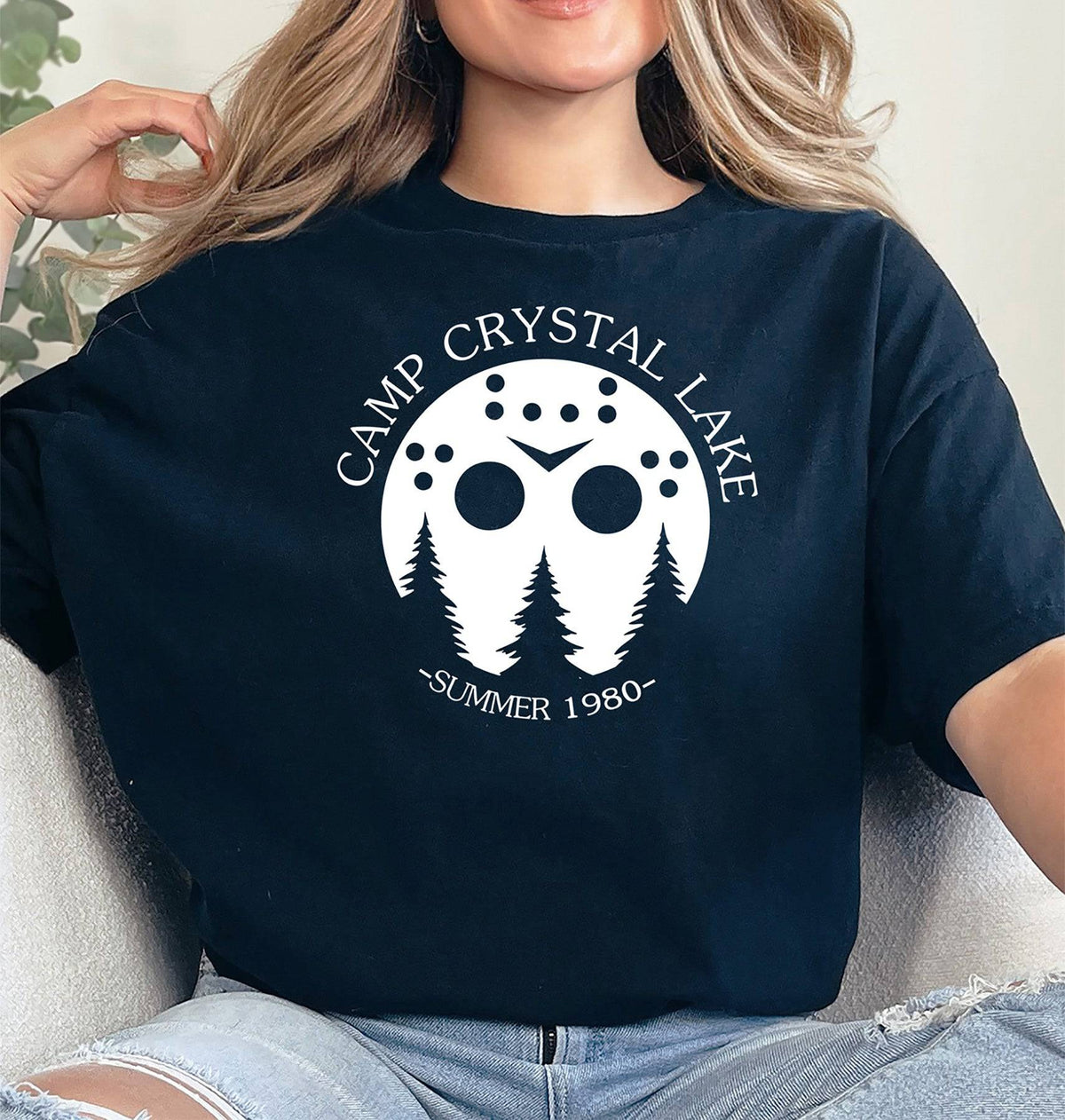 Woman wearing a black Camp Crystal Lake graphic T Shirt featuring a white hockey mask, pine trees, and Summer 1980 lettering.
