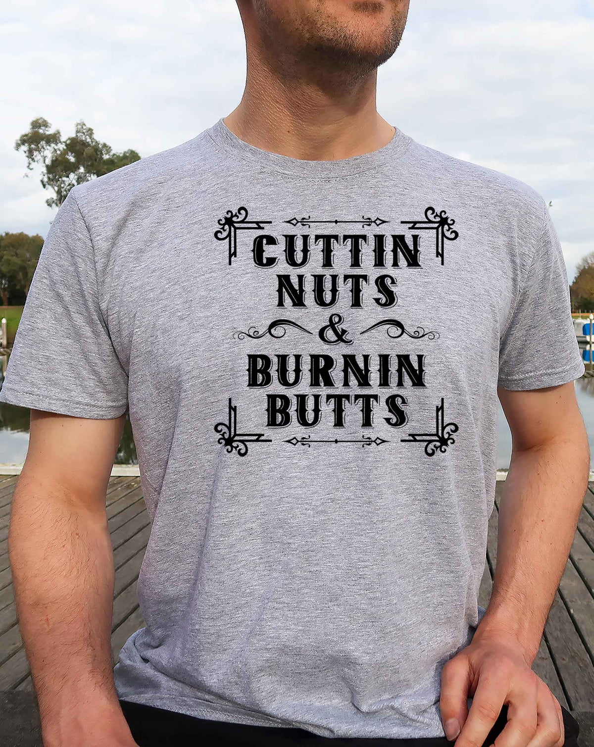 Man wearing a sport grey Cuttin Nuts & Burnin Butts graphic T Shirt featuring black vintage western lettering and decorative flourishes inspired by working cattle.