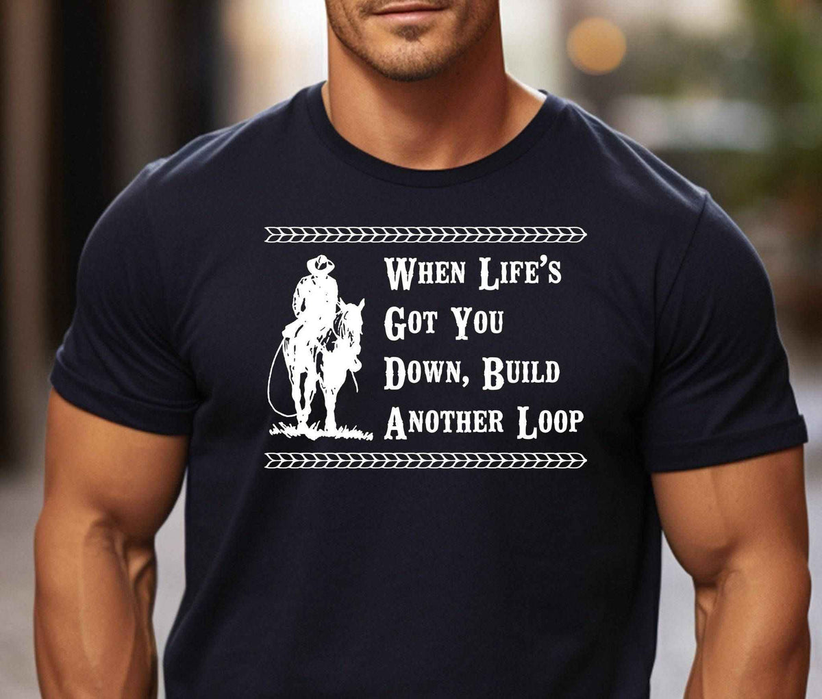 Man wearing a black western graphic T Shirt featuring a cowboy on horseback and the saying When Life's Got You Down, Build Another Loop.